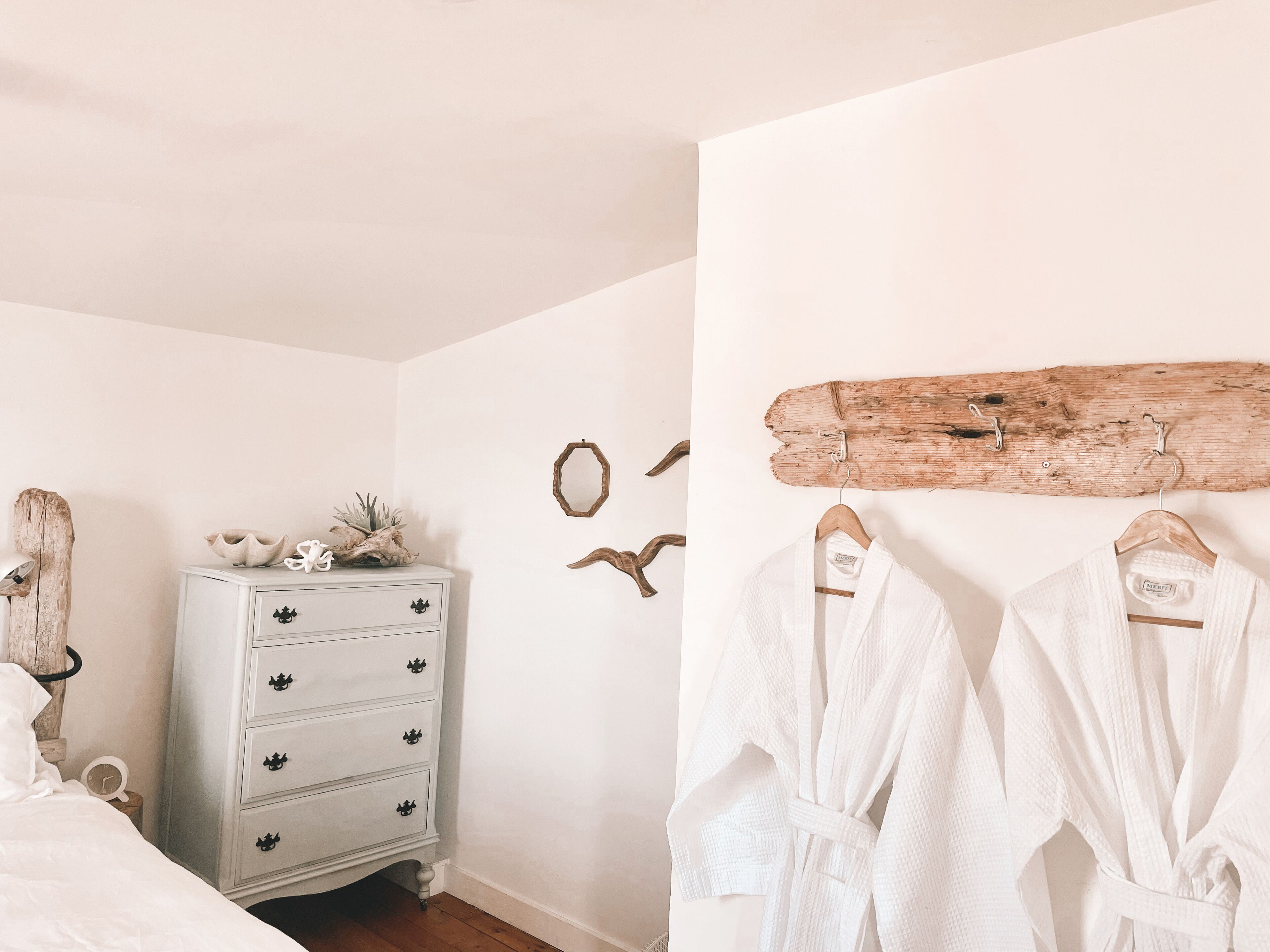 A bedroom with a white dresser, a wooden wall decor, and two white bathrobes hanging on hooks.
