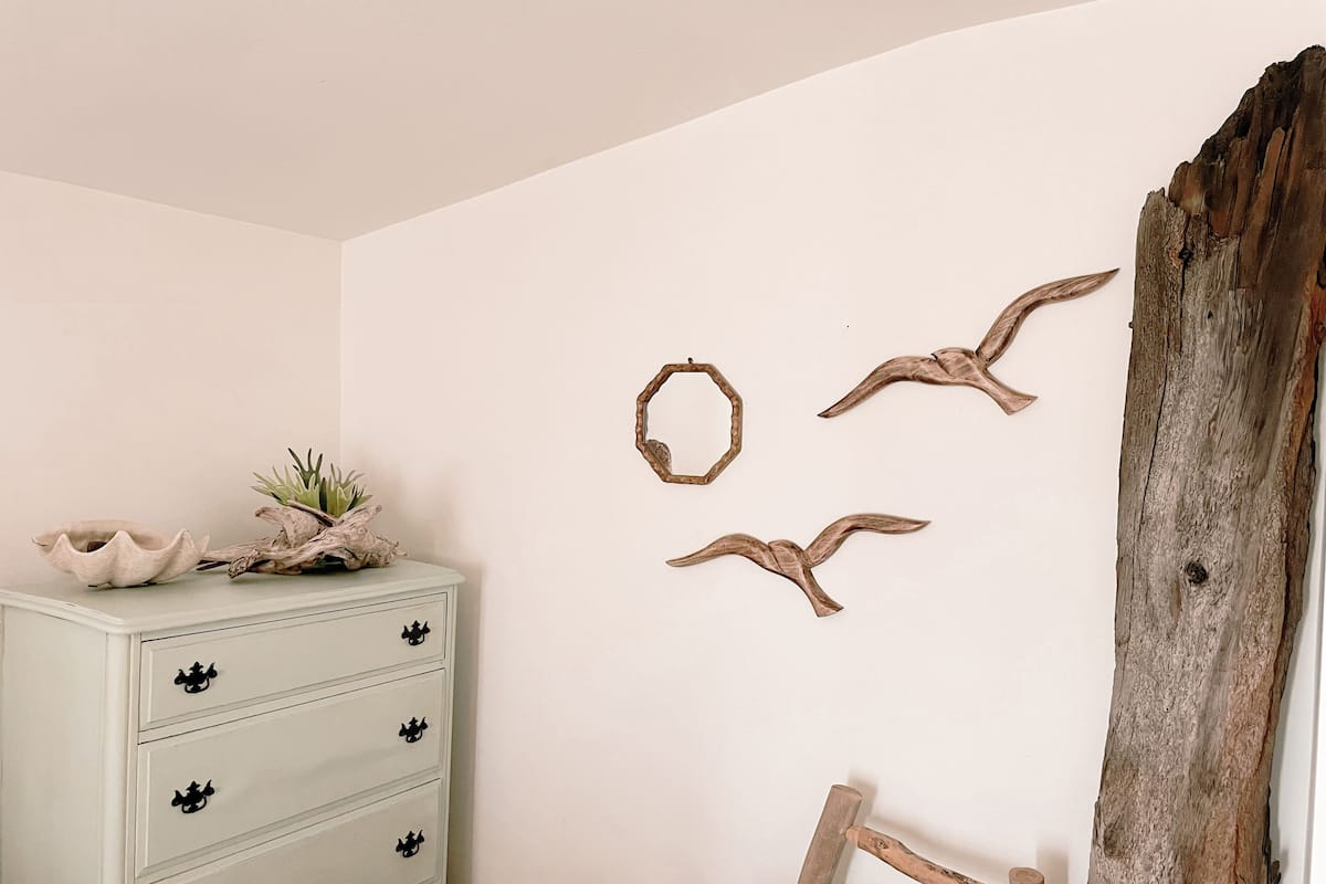 Ein Zimmer mit einem weißen Schminktisch, einem Holzstuhl und Wanddekorationen, darunter ein Vogel und ein runder Kranz.