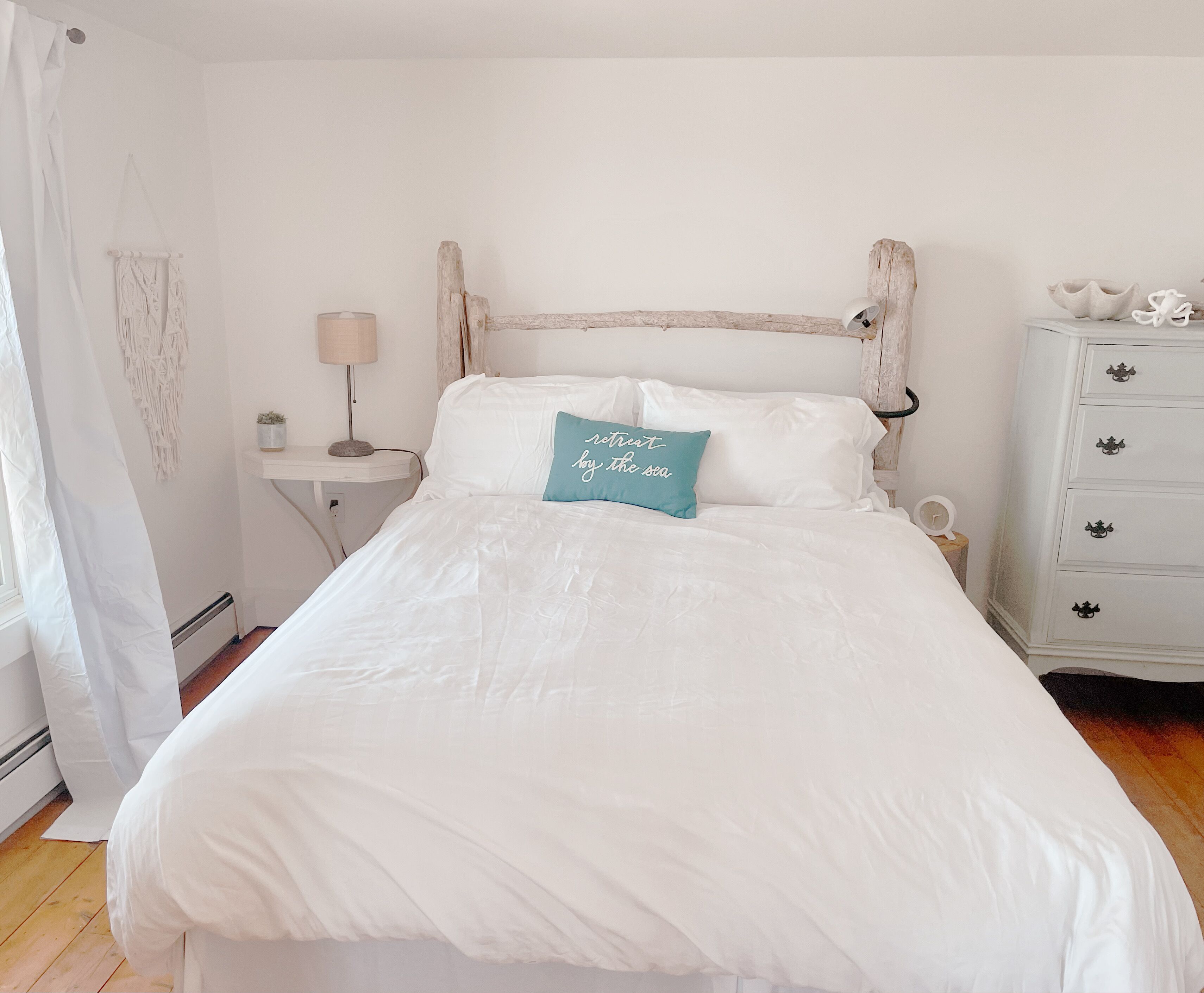 Un dormitorio con cama blanca, cabecera de madera, mesita de noche con lámpara y cómoda con cajones.