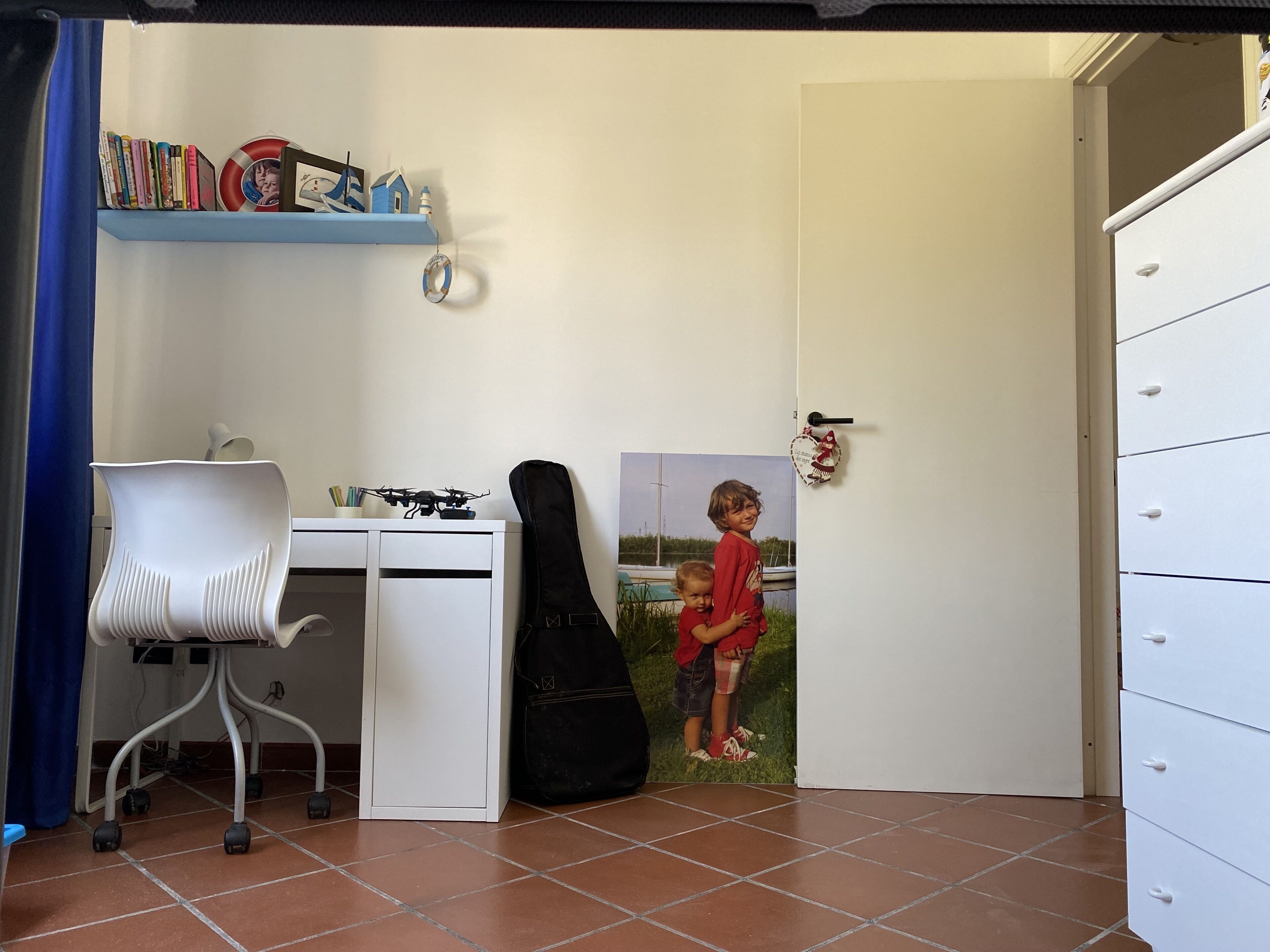 Quarto com uma escrivaninha branca, uma cadeira, um violão e uma porta com foto de uma criança.