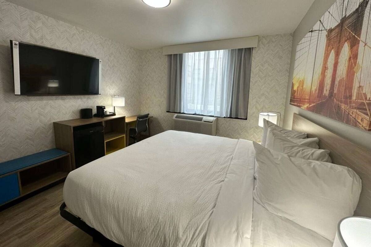 Habitación de hotel con una cama grande, un televisor de pantalla plana montado en la pared, un escritorio con una silla, una ventana con cortinas y un aire acondicionado de pared.