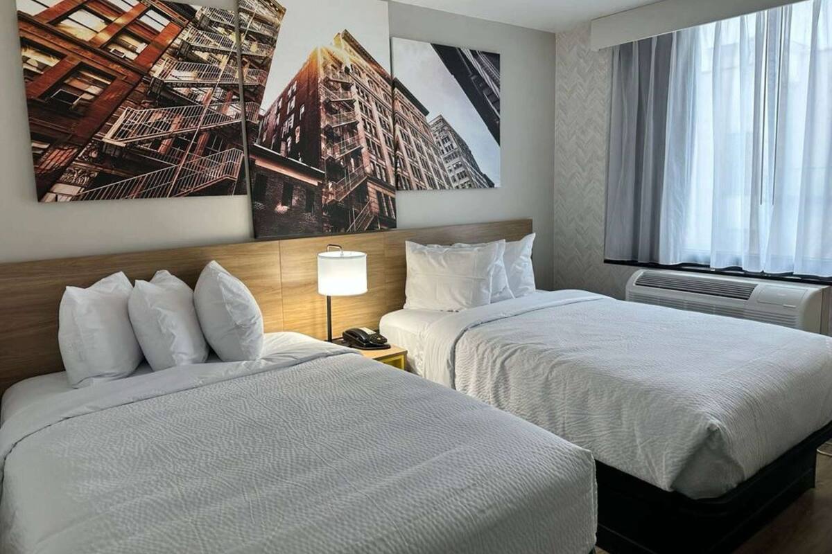 Habitación de hotel con dos camas, cabecero de madera, mesita de noche con lámpara y un ventanal con cortinas translúcidas.