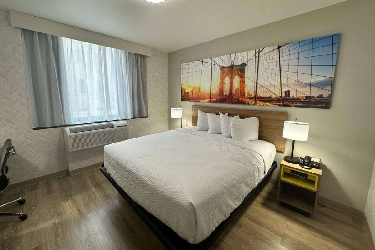 Habitación de hotel con una cama grande, una ventana con cortinas traslúcidas, un cuadro colgado en la pared que muestra un puente, una mesita de noche con una lámpara y una silla.