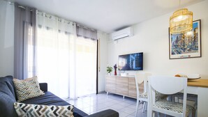 A modern living room with a sofa, a dining area with a white chair, a TV mounted on the wall, and a large window with curtains. - 5 min to Cala Crancs | Terrace & Pool (Salou)