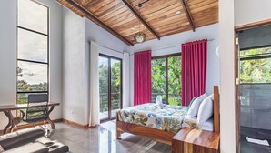 A modern bedroom with a wooden ceiling, a large bed with a floral bedspread, a desk with a chair, and a view of greenery through the windows.