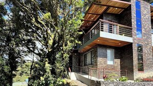 A modern two-story house with a stone facade, a wooden balcony, and a large tree in the foreground.
