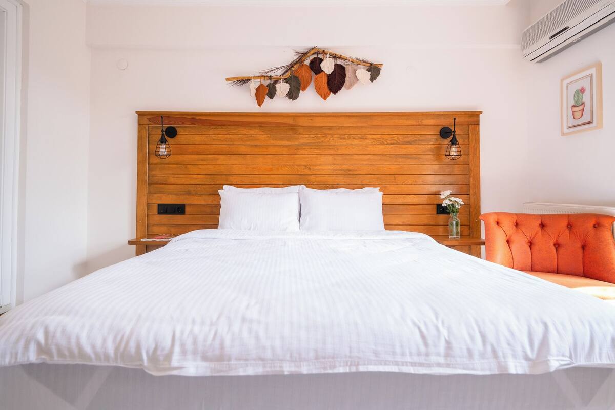 A bedroom with a wooden headboard, white bedding, a decorative wall hanging, and a small bedside table with a vase.