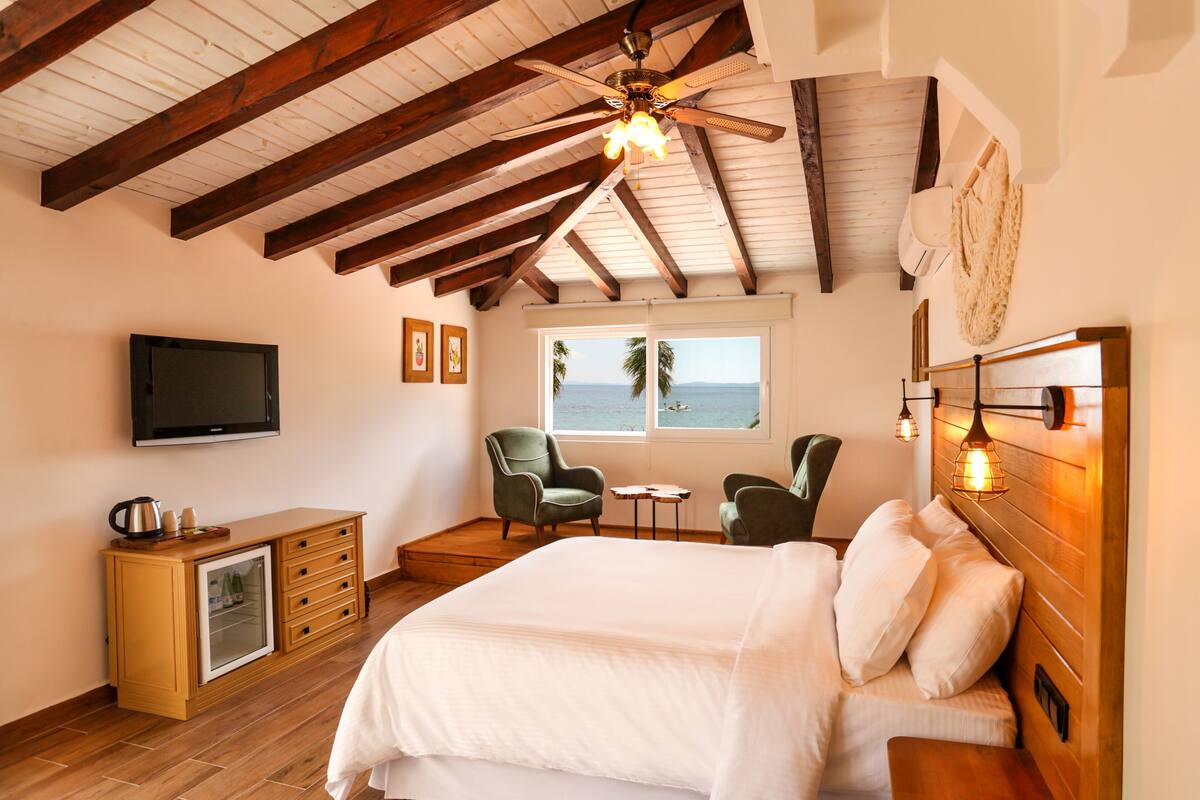 A bedroom with a wooden ceiling, a bed with white bedding, a television, a small table, two chairs, and a window with a view of the sea.