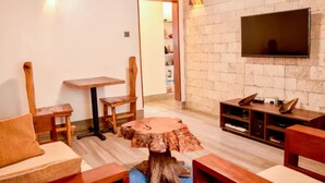 A cozy living room with a wooden coffee table, a TV mounted on a brick wall, and a small dining table. - Secure, 2 bedroom house in a quiet neighborhood. (Nairobi County)