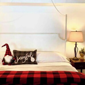 A neatly made bed with a red and black checkered blanket, a pillow with a gnome and the word "Joyful," a lamp on a bedside table, and a white wall.
