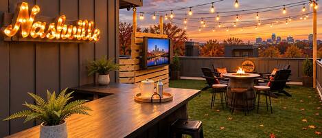 An outdoor patio with a wooden table, chairs, a TV screen, and string lights.