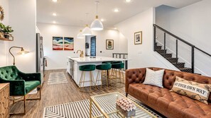 A modern living room with a brown leather sofa, green velvet armchair, and a dining area with a white island and green bar stools. - Johnny & June House • Walk to Bars & Dining (Nashville)