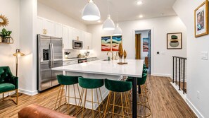 A modern kitchen with white cabinets, stainless steel appliances, a central island with bar stools, and a staircase with a railing. - Johnny & June House • Walk to Bars & Dining (Nashville)