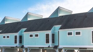 A row of light blue beach houses with dark roofs, each featuring a small porch and a sign indicating the name of the beach house. - Easy Walk to Beach, Boardwalk, & Lake! King Beds! (Carolina Beach)