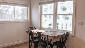 A dining area with a wooden table and four chairs, large windows with blinds, and a view of a white fence. - Easy Walk to Beach, Boardwalk, & Lake! King Beds! (Carolina Beach)