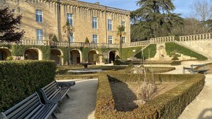 Un giardino formale con panchine, una fontana e un edificio storico con finestre ad arco e facciata in pietra.