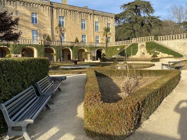 Un jardín formal con una fuente, bancos y un edificio clásico con ventanas arqueadas.