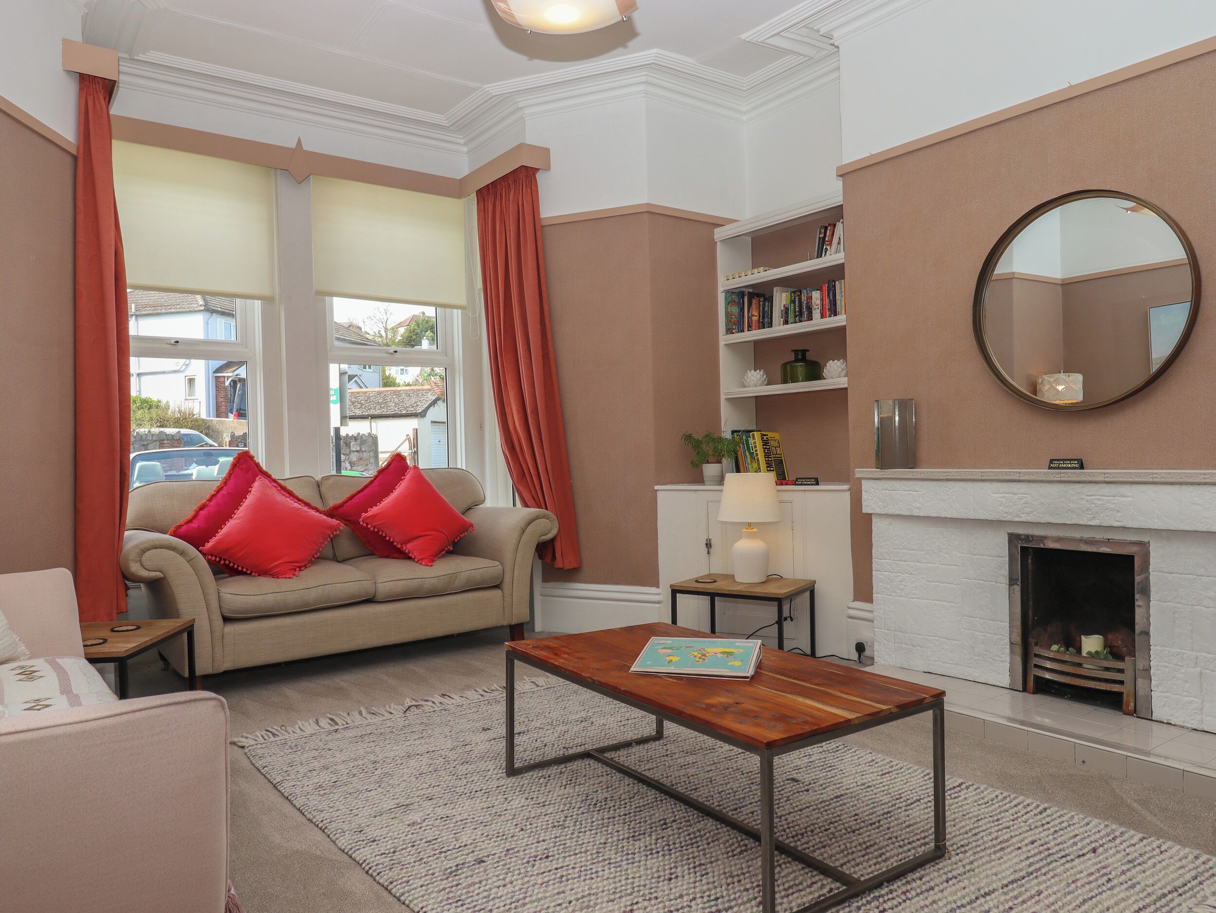 A living room with a beige sofa, red cushions, a wooden coffee table, a bookshelf, a fireplace, and a large mirror.