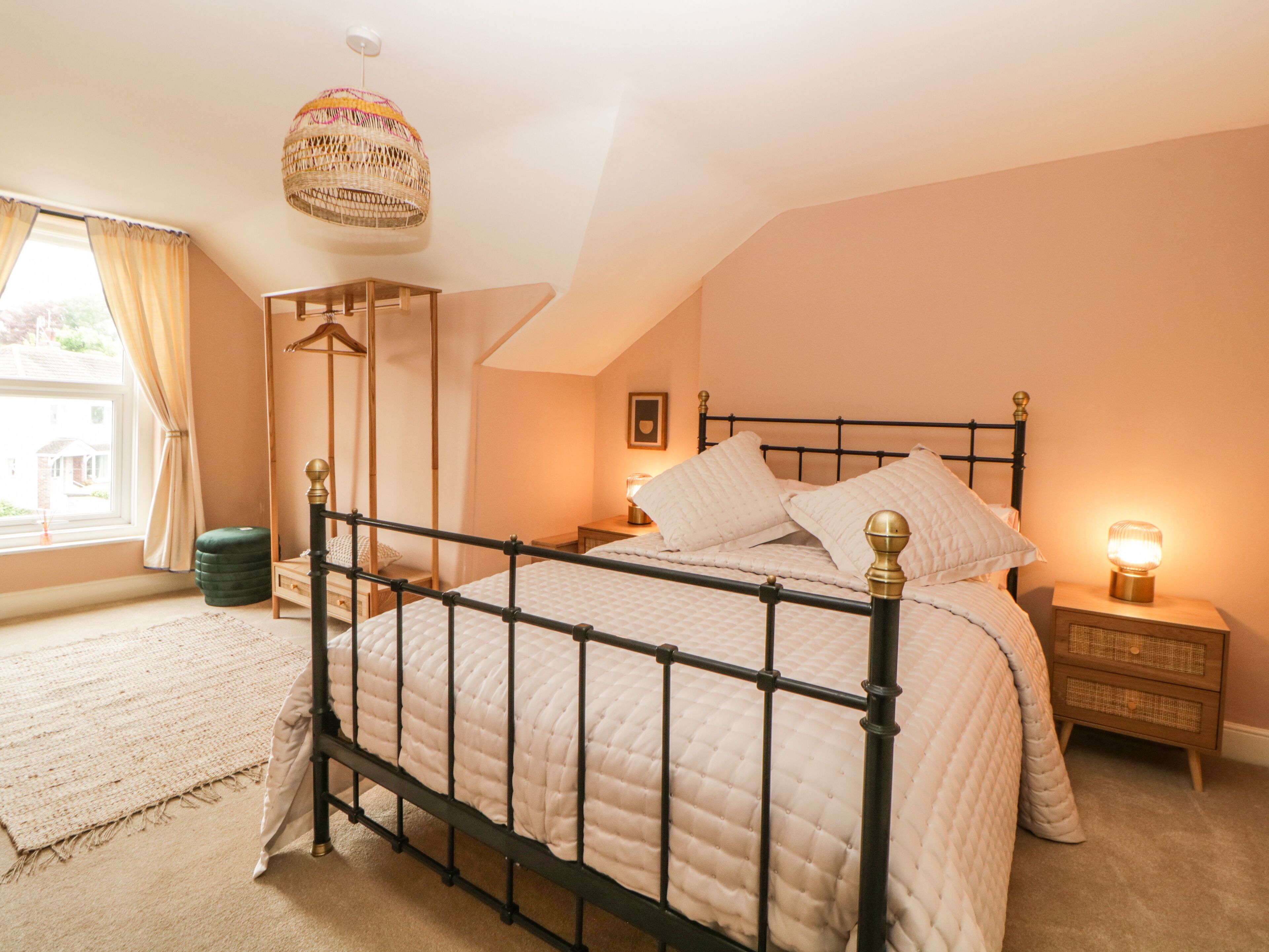 A bedroom with a four-poster bed, a hanging light fixture, a bedside table with a lamp, and a view of the outside through a window with curtains.