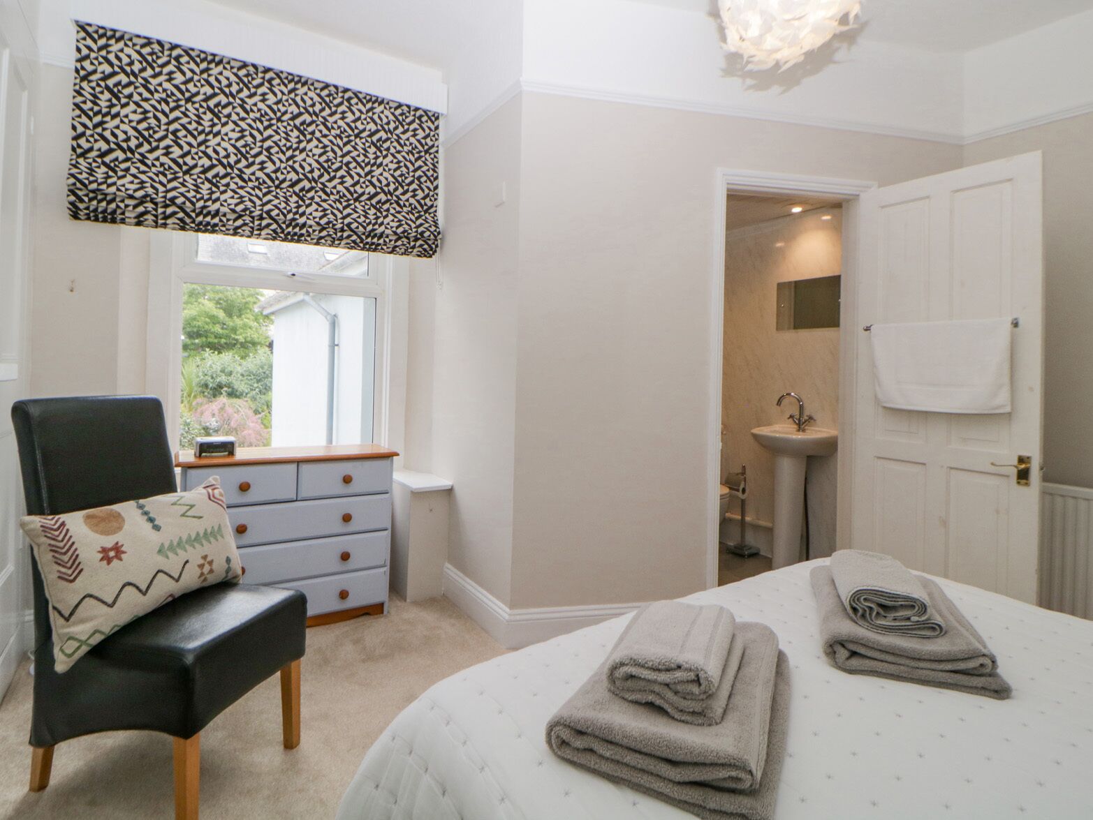 A bedroom with a bed, a chair, a dresser, a window, and a bathroom visible through an open door.