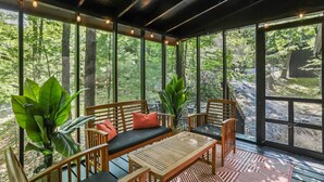 Un área de estar al aire libre con techo, mobiliario de madera, ventanas amplias y plantas en macetas.