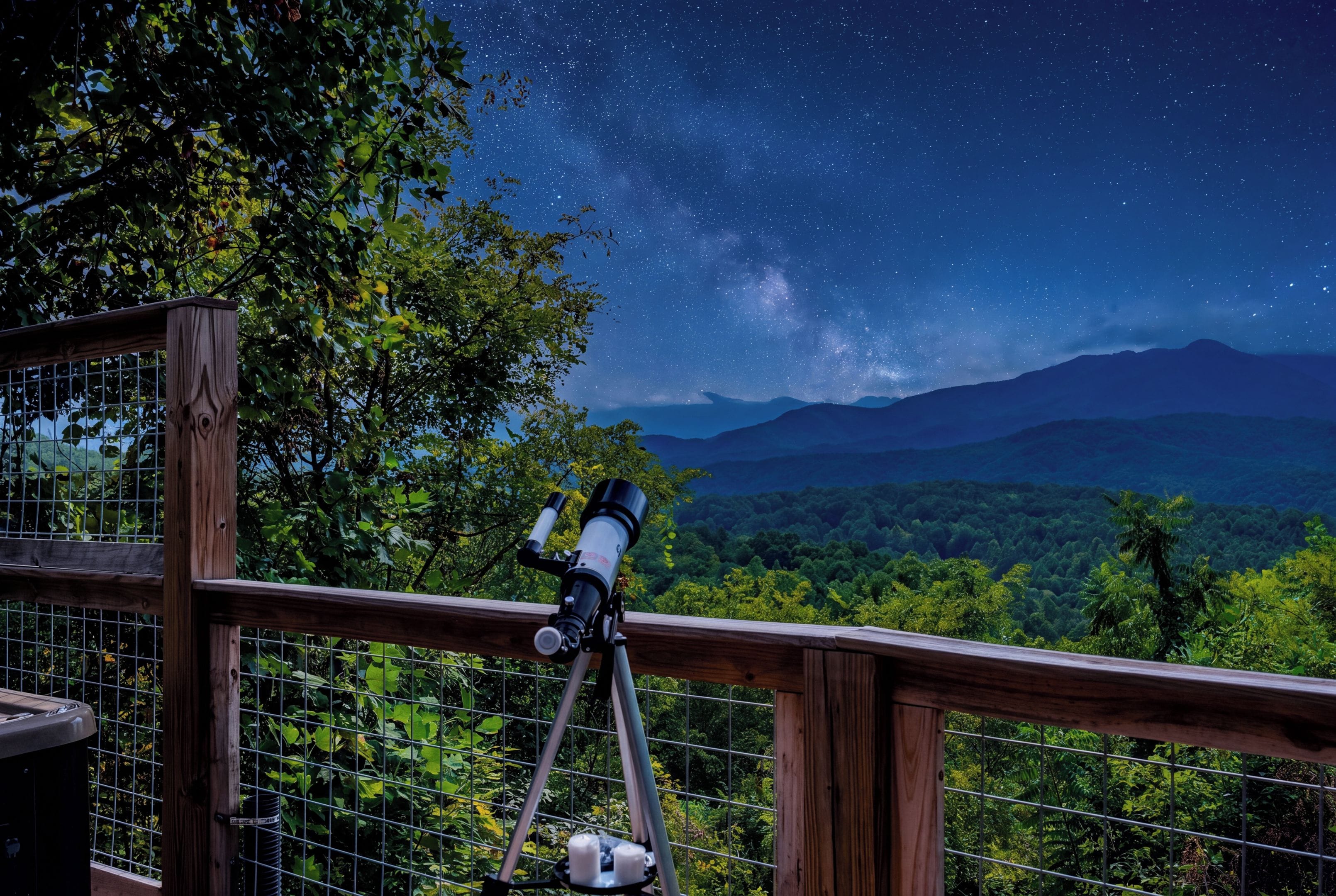 A telescope on a wooden deck overlooking a mountainous landscape with a starry night sky.