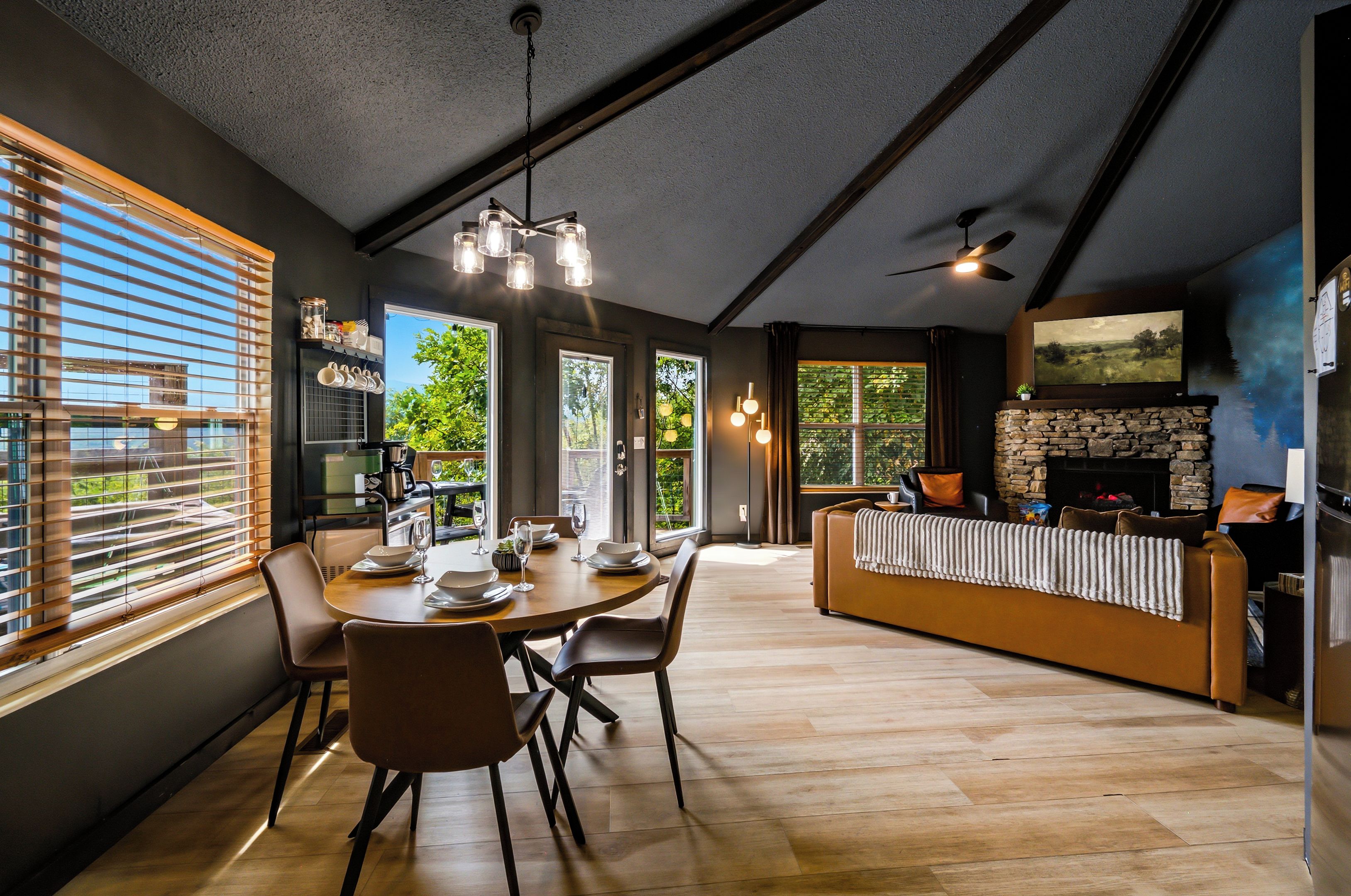 A modern dining area with a wooden table and chairs, a kitchen with a stone fireplace, and large windows with blinds.