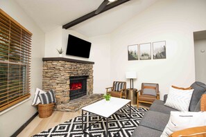 A living room with a stone fireplace, a flat-screen TV mounted above it, and a sofa with patterned cushions.