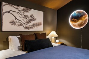 A bedroom with a bed, pillows, a nightstand with a lamp, and a wall with a tree artwork and a spherical celestial artwork.