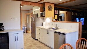 A compact kitchen with white cabinets, a microwave, a sink, and a dishwasher. There is a wooden ceiling and a clock on the wall. - Large cabin on rural farm.  Private, relaxing, close to Stamp Falls Park. (Port Alberni)