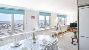 A dining area with a table set for four, a bottle of wine, and a view of the sea and parked vehicles through large windows.