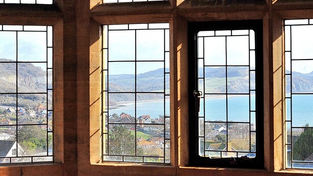 A room with multiple windows featuring a grid pattern, offering views of a coastal landscape with hills and a body of water. - 3 Coram Tower (Lyme Regis)