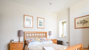 A bedroom with a wooden bed, two bedside tables, a nightstand, a window with blinds, and two framed pictures on the wall. - 3 Coram Tower (Lyme Regis)