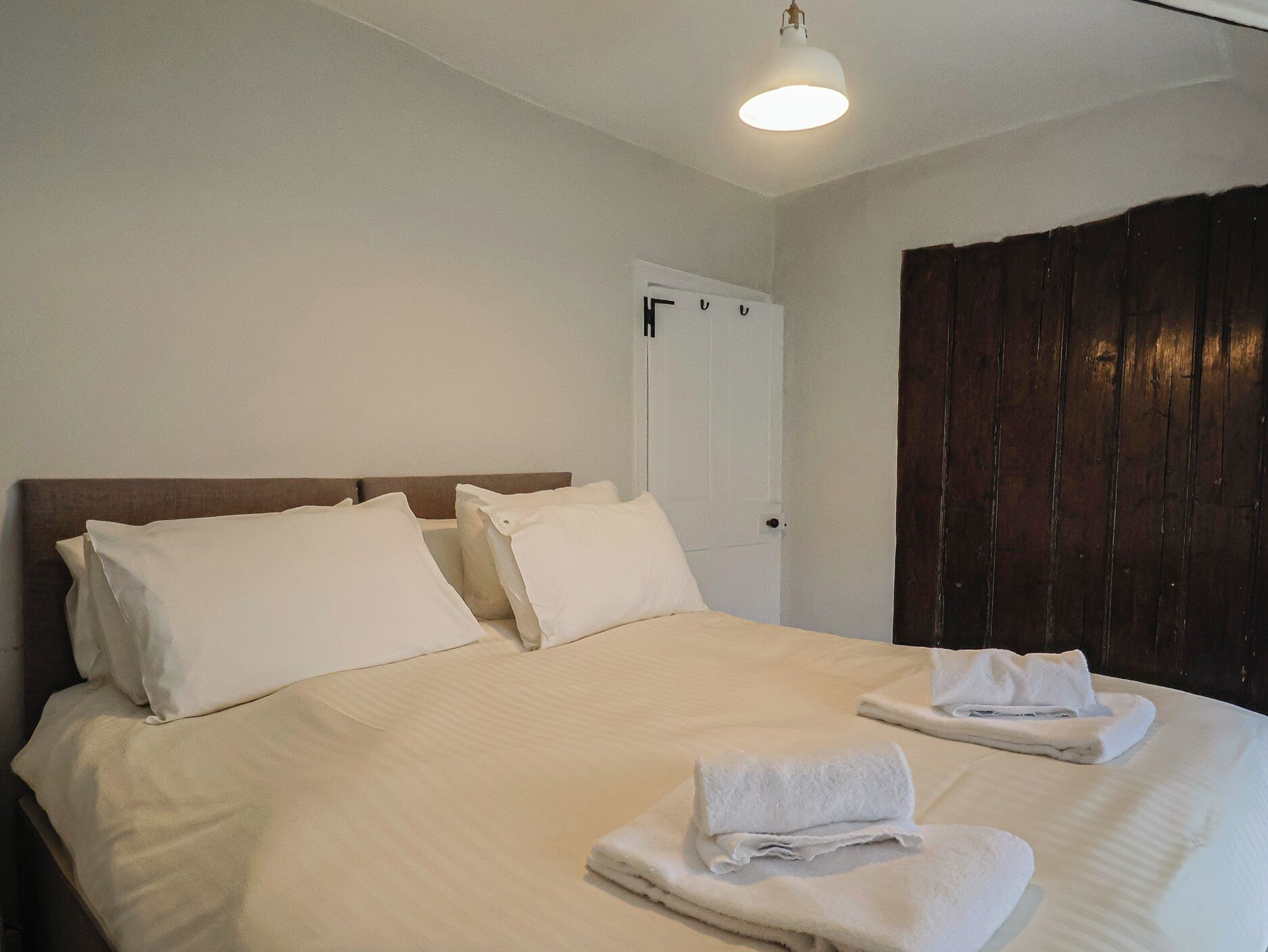 A neatly made bed with white linens and two pillows, a wooden headboard, a white door, and a dark wooden panel on the wall.