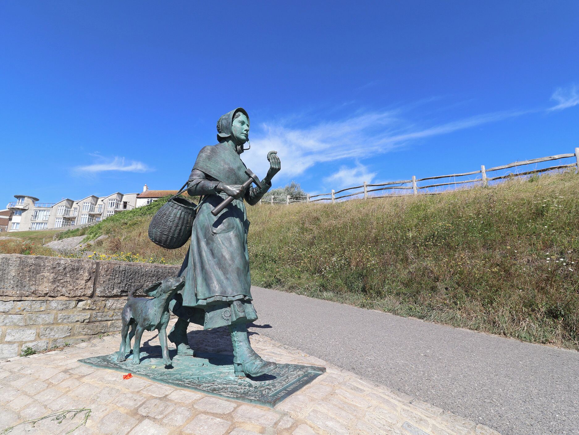 A statue of a woman holding a basket and a stick, standing on a paved area with a stone wall and a wooden fence in the background.