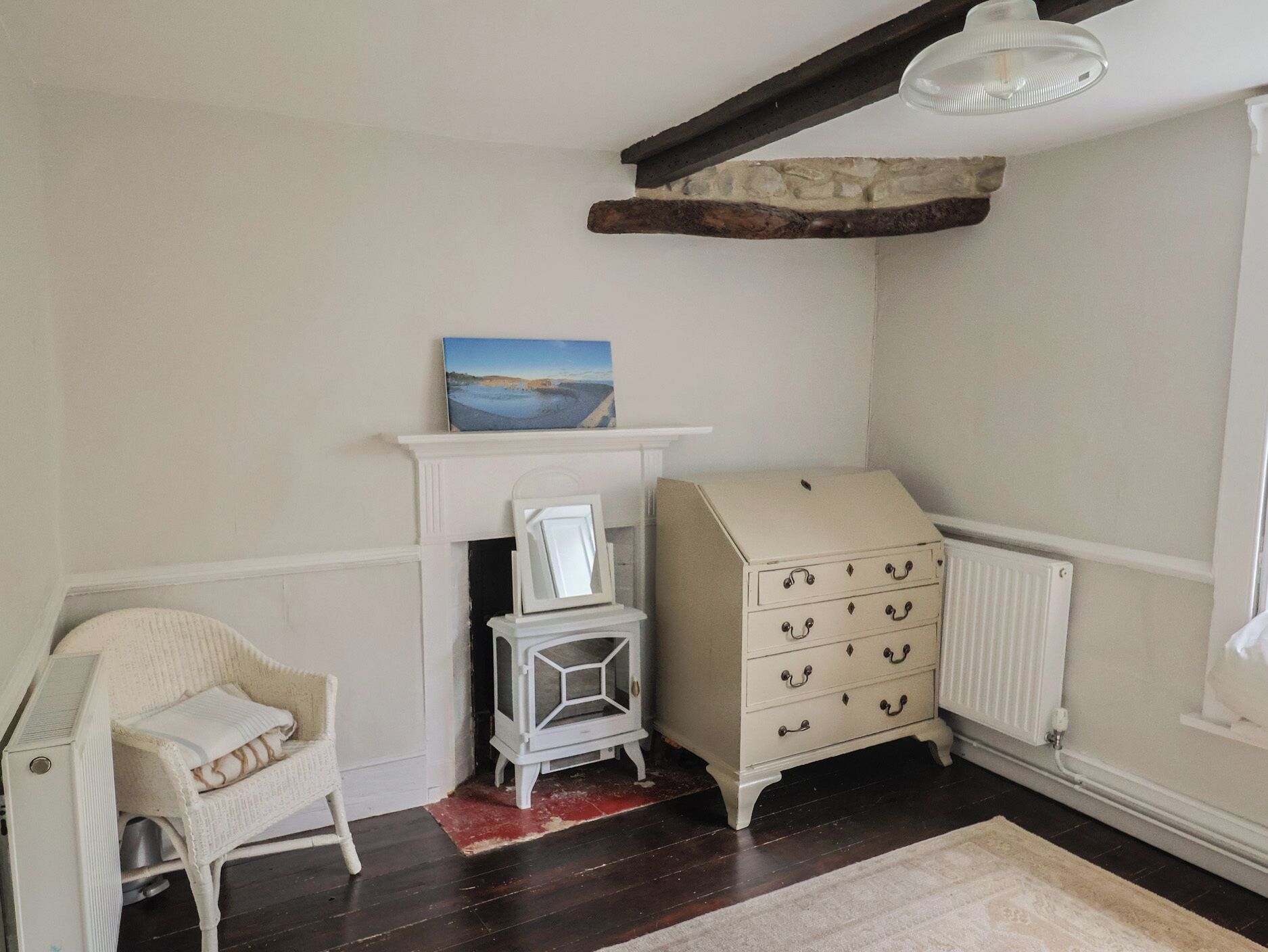 Een kamer met een witte stoel, een kaptafel, een klein tafeltje, een schilderij boven de schoorsteenmantel en een radiator.