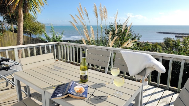 Une terrasse en bois avec une table dressée pour deux personnes, agrémentée d’une bouteille de vin, de deux verres, d’un livre et d’une paire de lunettes.