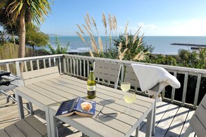 A wooden deck with a table set for two, featuring a bottle of wine, two glasses, a book, and a pair of glasses. - The Cabin (Lyme Regis)
