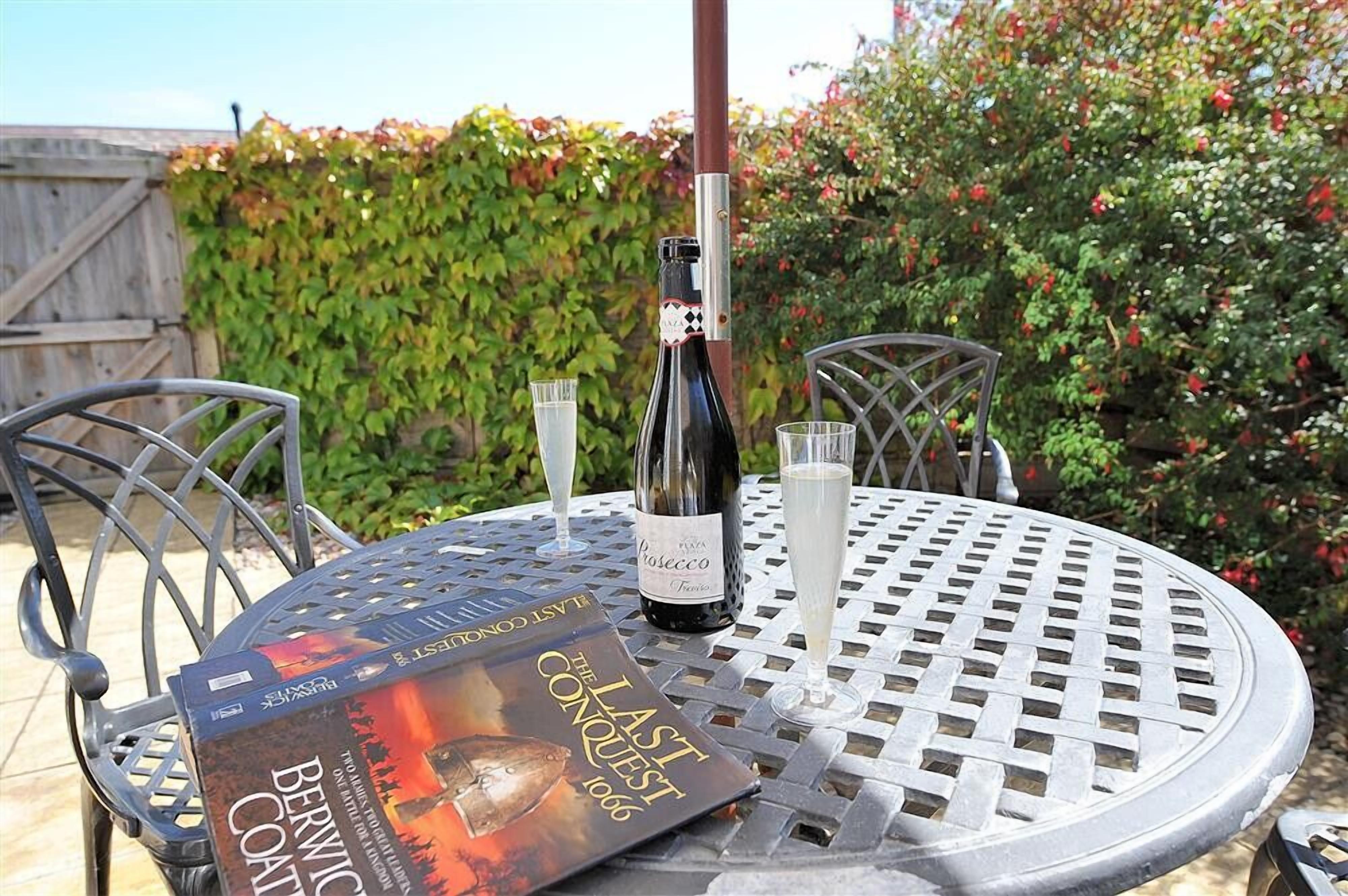 A round metal table with a bottle of wine and two glasses, a book titled "The Last Conquest 1066" on it, and a metal chair.