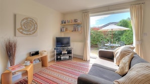 A living room with a sofa, TV, bookshelf, and a view of a garden through a sliding door. - 1 Coppers Knapp (Lyme Regis)