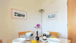 A dining table set with white bowls, plates, and cutlery, a vase with a pink flower, a glass of orange juice, and a jar of jam. - 1 Coppers Knapp (Lyme Regis)