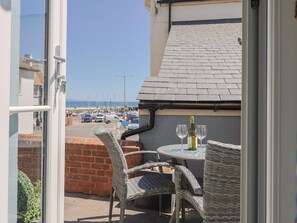 Un balcón con sillas de mimbre y una mesa, con vista a un puerto deportivo con barcos y un cielo despejado.