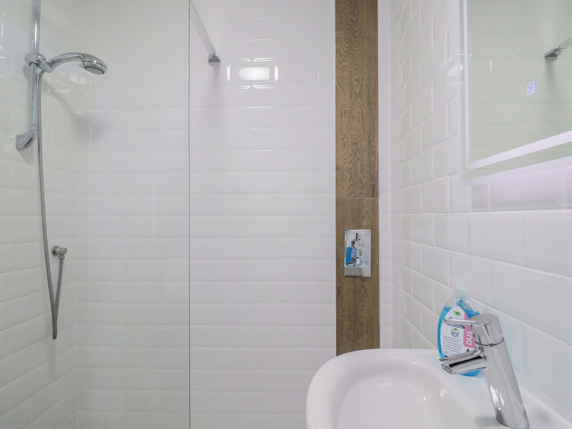 A bathroom with white tiled walls, a wooden accent wall, a shower with a handheld showerhead, a sink with a faucet, and a soap dispenser.