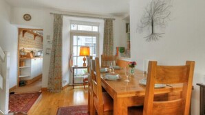 A dining area with a wooden table set for four, flanked by chairs. A window with curtains is visible in the background, and a lamp is on the table. - Windwhistle (Lyme Regis)