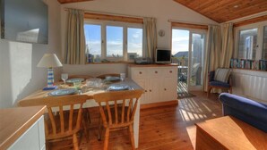 Una cocina acogedora con techo de madera y ventanas amplias, una mesa de comedor con platos y vasos, y un sofá azul en la sala.
