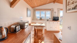 Una cocina con vigas de madera, una zona de comedor con sillas, una mesa, un hervidor, un tostador y un televisor.