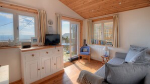 Un cuarto acogedor con techo de madera, un sofá, un televisor sobre un mueble de madera, y vista al mar a través de amplias ventanas.