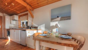 Una cocina de madera con armarios blancos, una mesa de comedor preparada para cuatro comensales y un cuadro con una escena de playa en la pared.