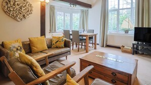 A cozy living room with a sofa, coffee table, and dining table. There are windows with curtains, a television, and a decorative wall piece. - Maries (Lyme Regis)