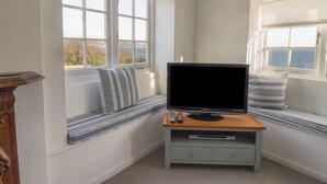 A room with a television on a wooden stand, a remote control, and a DVD player on a shelf. There are two windows with blinds, and a bench with striped cushions. - 4 East Cliff (Lyme Regis)